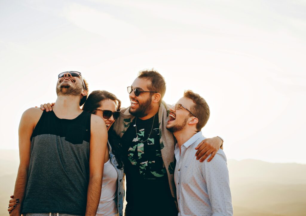 Four joyful young adults, appearing to be friends, share a laugh outdoors with bright sunlight behind them.