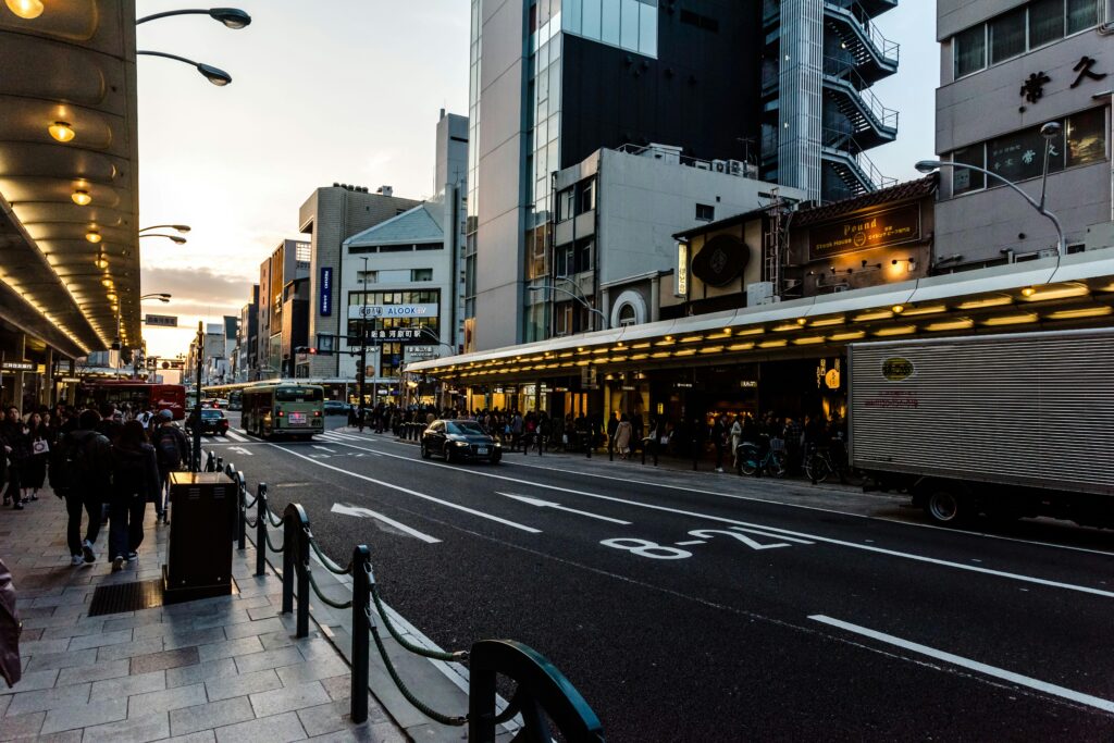 An evening street scene in Kyoto, Japan, with a bustling sidewalk, cars on the road, and buildings illuminated by lights.