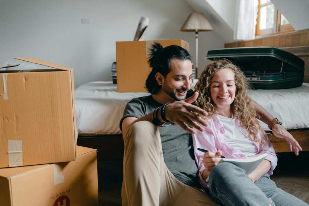 A smiling couple sits on the floor surrounded by moving boxes, as the woman writes in a notebook, suggesting they are settling into a new home.