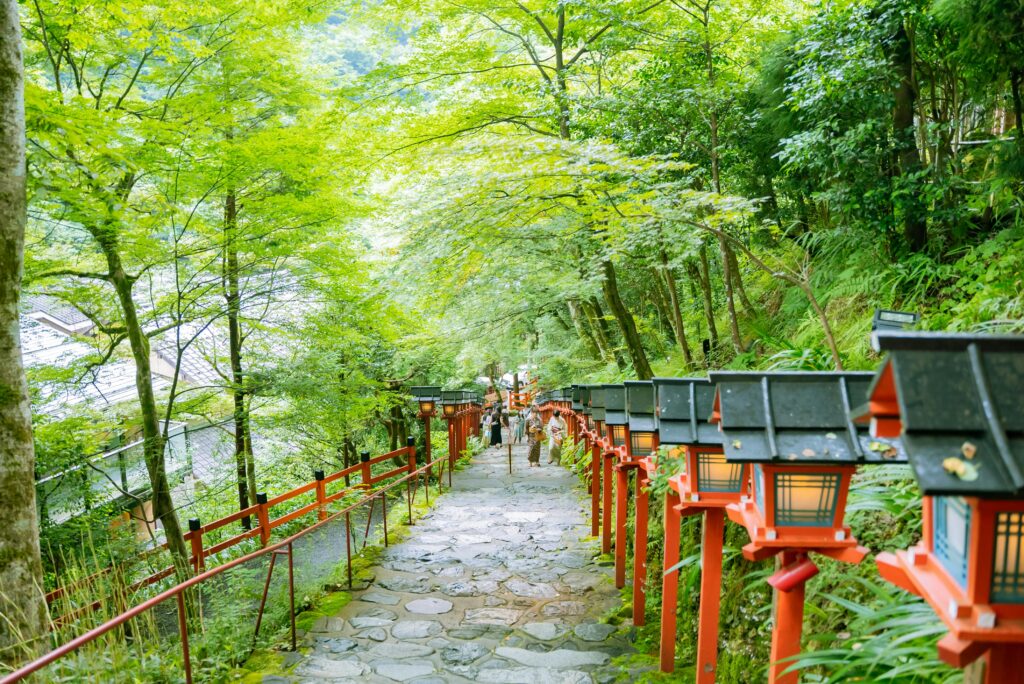 A scenic stone pathway lined with traditional red Japanese lanterns and lush green trees leads up a mountain.